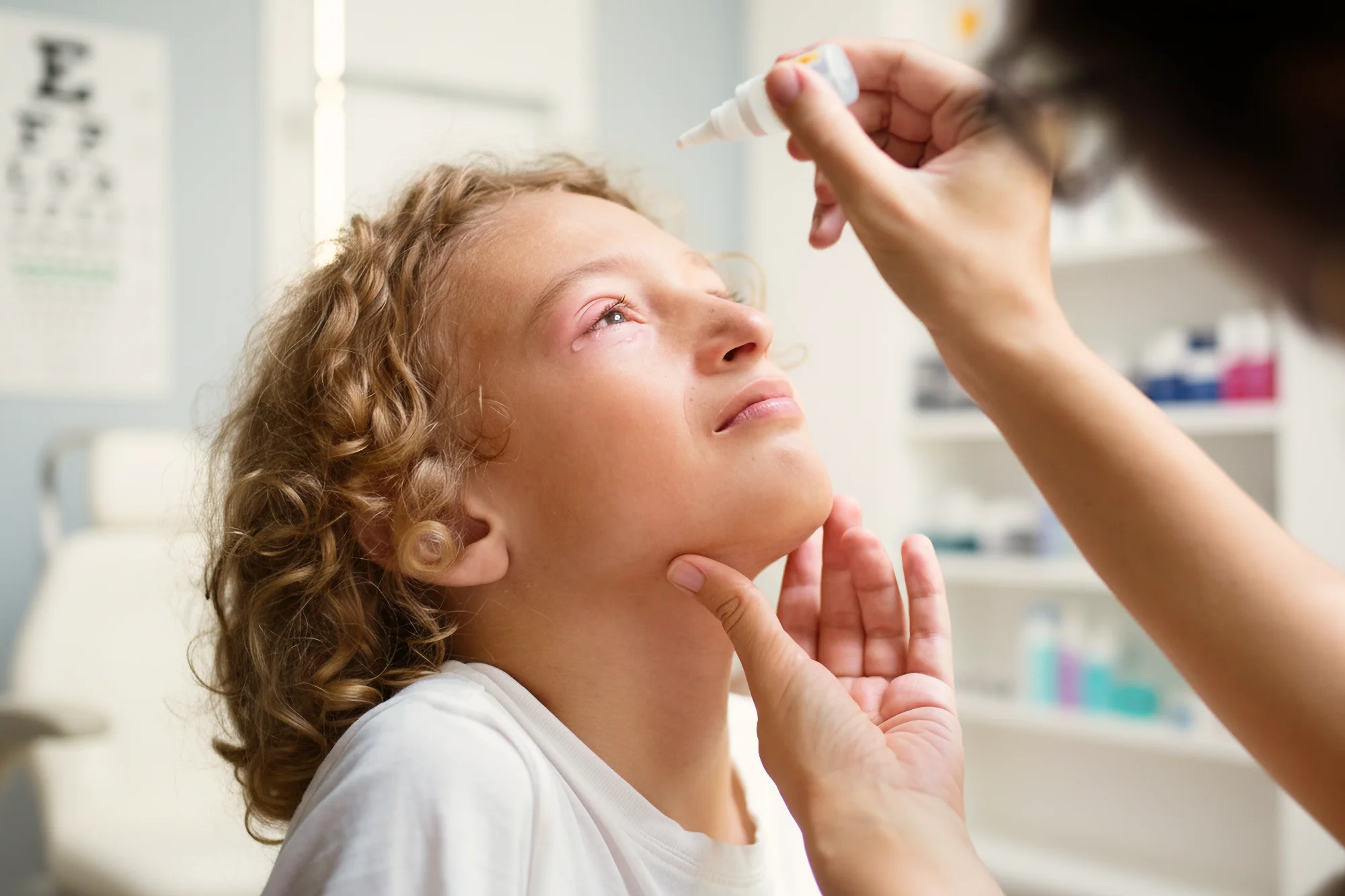 A person administers eye drops into the irritated eye of a young patient with curly blonde hair in an eye clinic. The patient holds still and looks up.