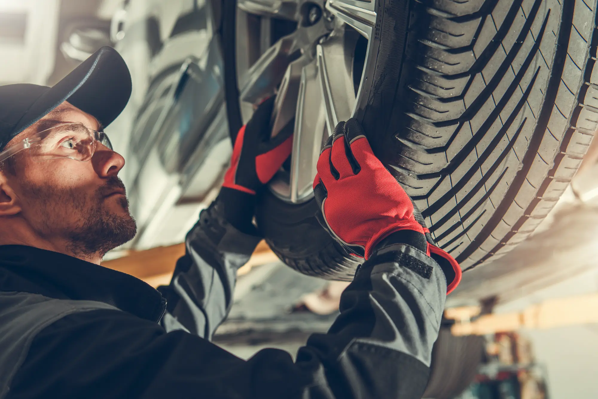 A male mechanic with a focused expression wearing safety glasses and red gloves installs a new tire onto a lifted vehicle in a garage.