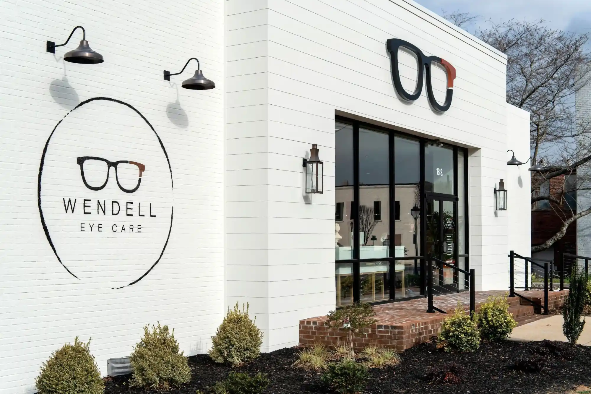 The contemporary white exterior of the Wendell Eye Care office, featuring a prominent logo of black and orange eyeglasses above the main glass entrance.