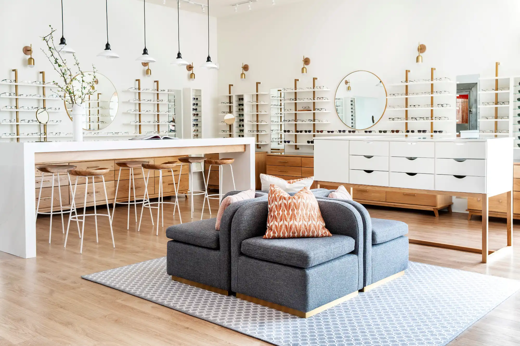A bright, modern optical shop interior featuring walls lined with white shelves displaying eyeglasses. A central gray seating area sits on a rug near a white counter with wooden stools.
