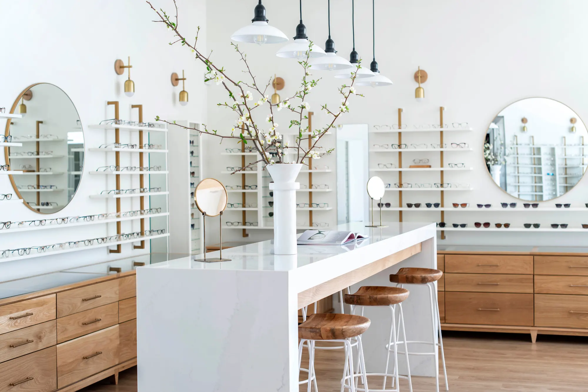 A bright, modern optician’s retail space with white walls and shelves displaying glasses. A central white counter has wood stools, a vase of flowering branches, and round gold mirrors nearby.