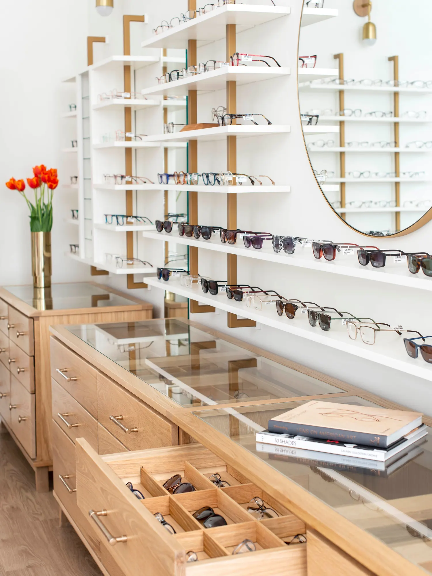 A modern optical shop interior featuring rows of eyeglasses and sunglasses displayed on white floating shelves above light wood cabinetry.