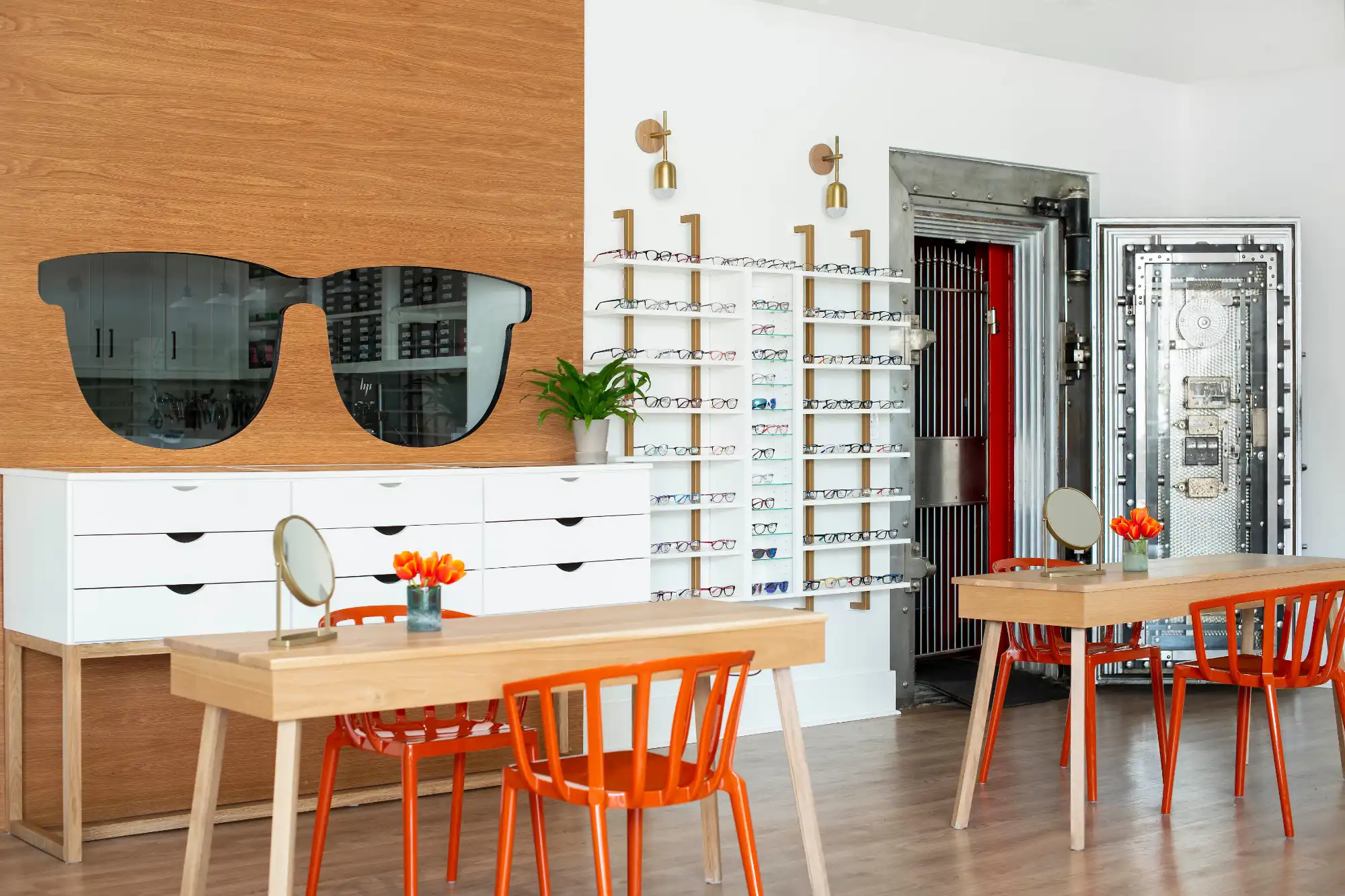 A modern optometrist office showing eyeglass displays, bright orange chairs, and two wooden tables. The unique decor includes a sunglasses mirror and a large, open metal bank vault door.