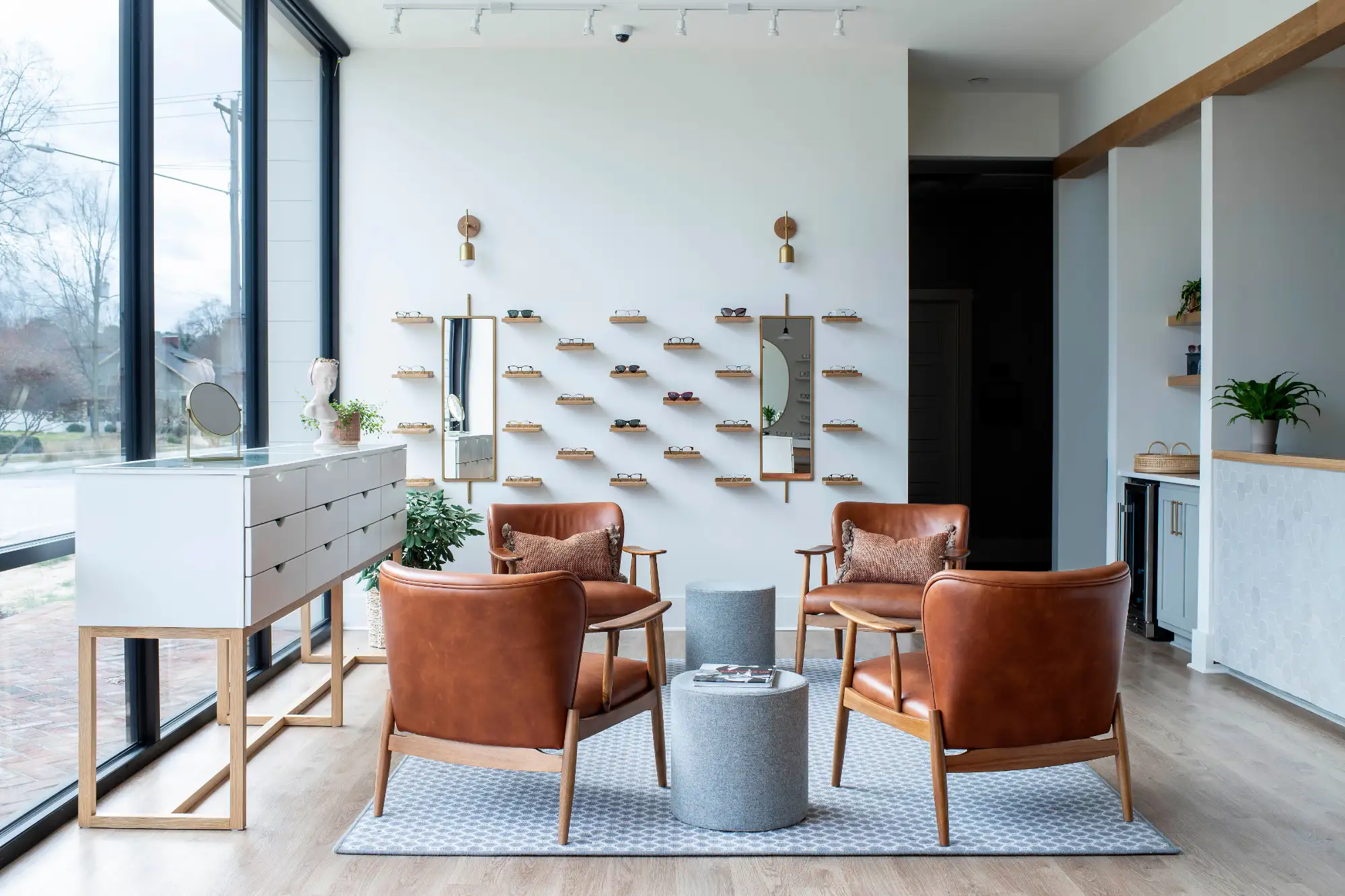An inviting optical shop waiting room with four brown leather chairs and small tables. Prescription glasses are displayed neatly on the white feature wall.