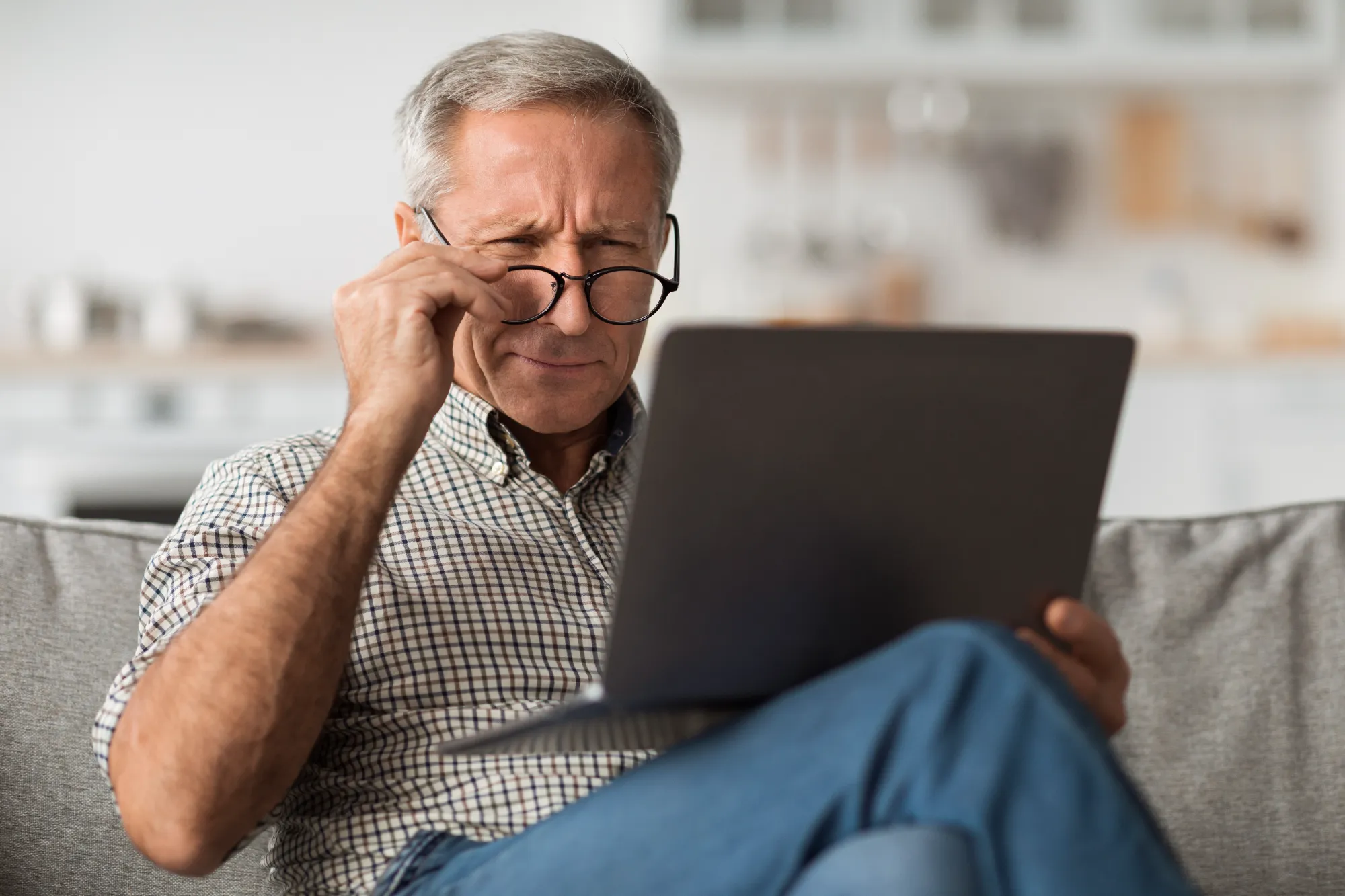 An older man with gray hair squints intensely and adjusts his glasses while trying to read information on a laptop screen.