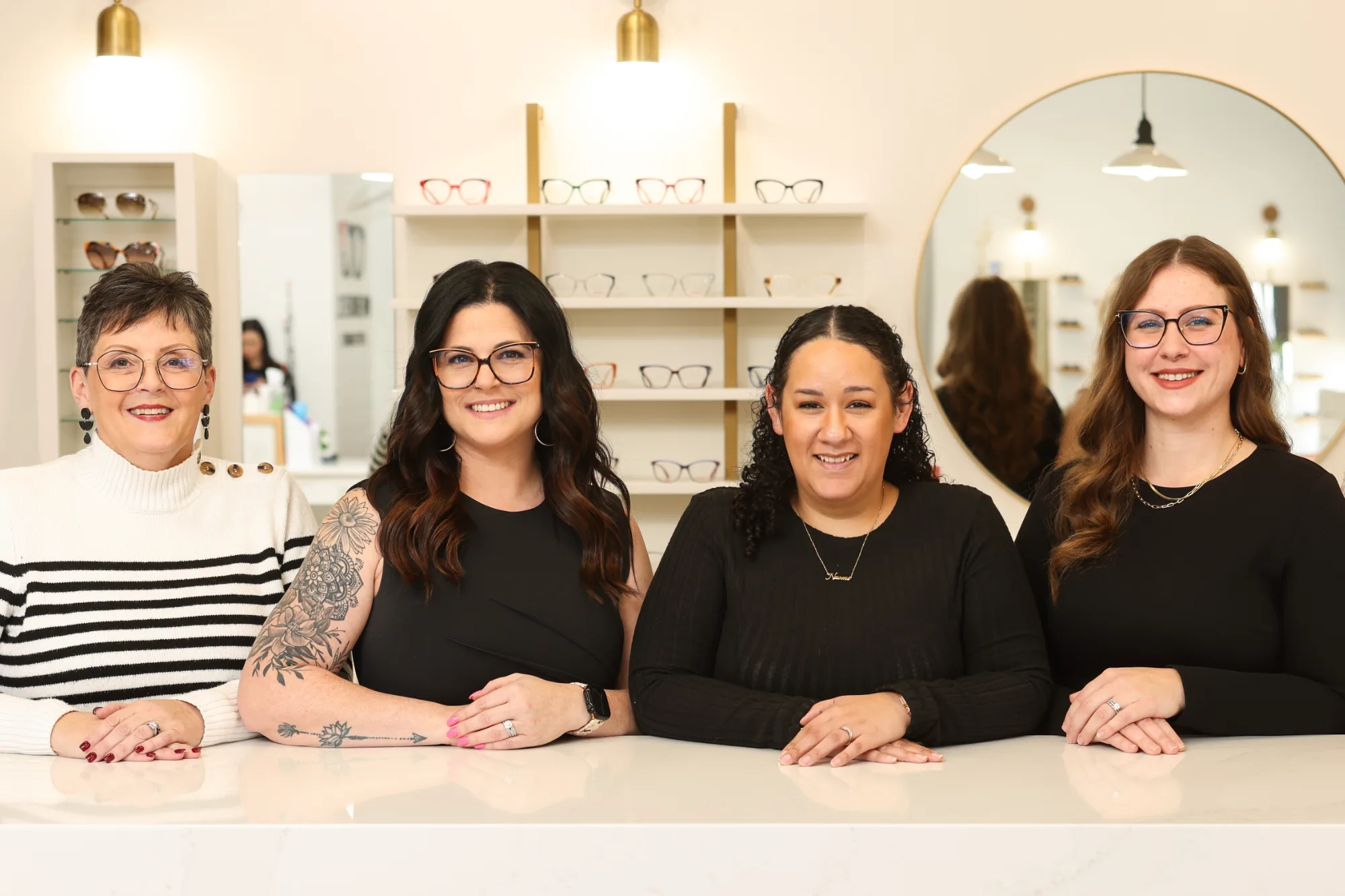 Four smiling women, the optical team, stand behind a white counter in the optometrist office, with various eyeglasses displayed on shelves behind them.