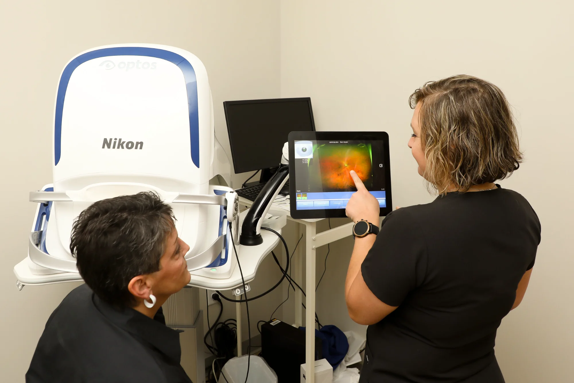 An eye care technician points to a patient's optomap scan on a monitor, explaining the results to the seated client in the examination room.