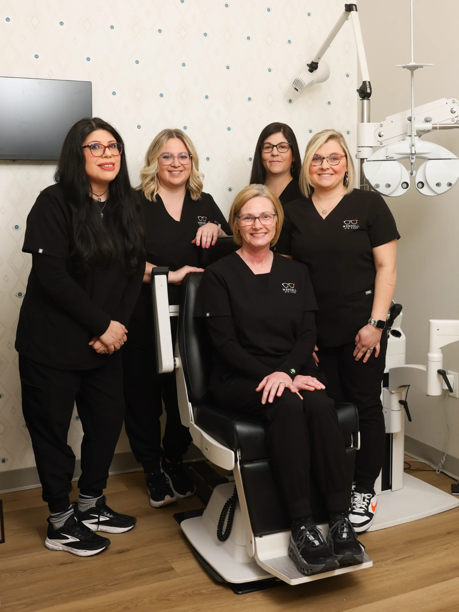 Five smiling optometric technicians from Wendell Eye Care, wearing black scrubs, pose next to an eye examination chair and phoropter.