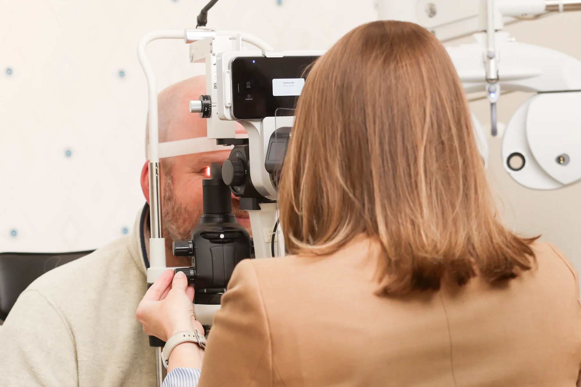Optometrist Dr. Holly Bennett examines a patient's eyes using a slit lamp machine. The doctor adjusts the dial while the patient looks into the lens.