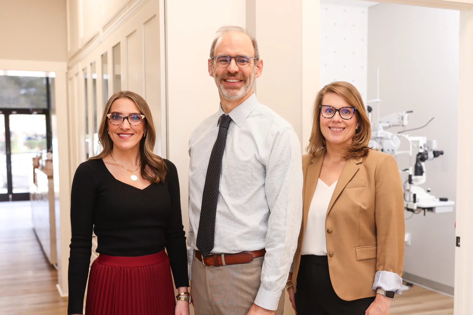 Three optometrists, two women and one man, stand together smiling in a brightly lit hallway of an eye clinic, with specialized eye examination equipment visible behind them.