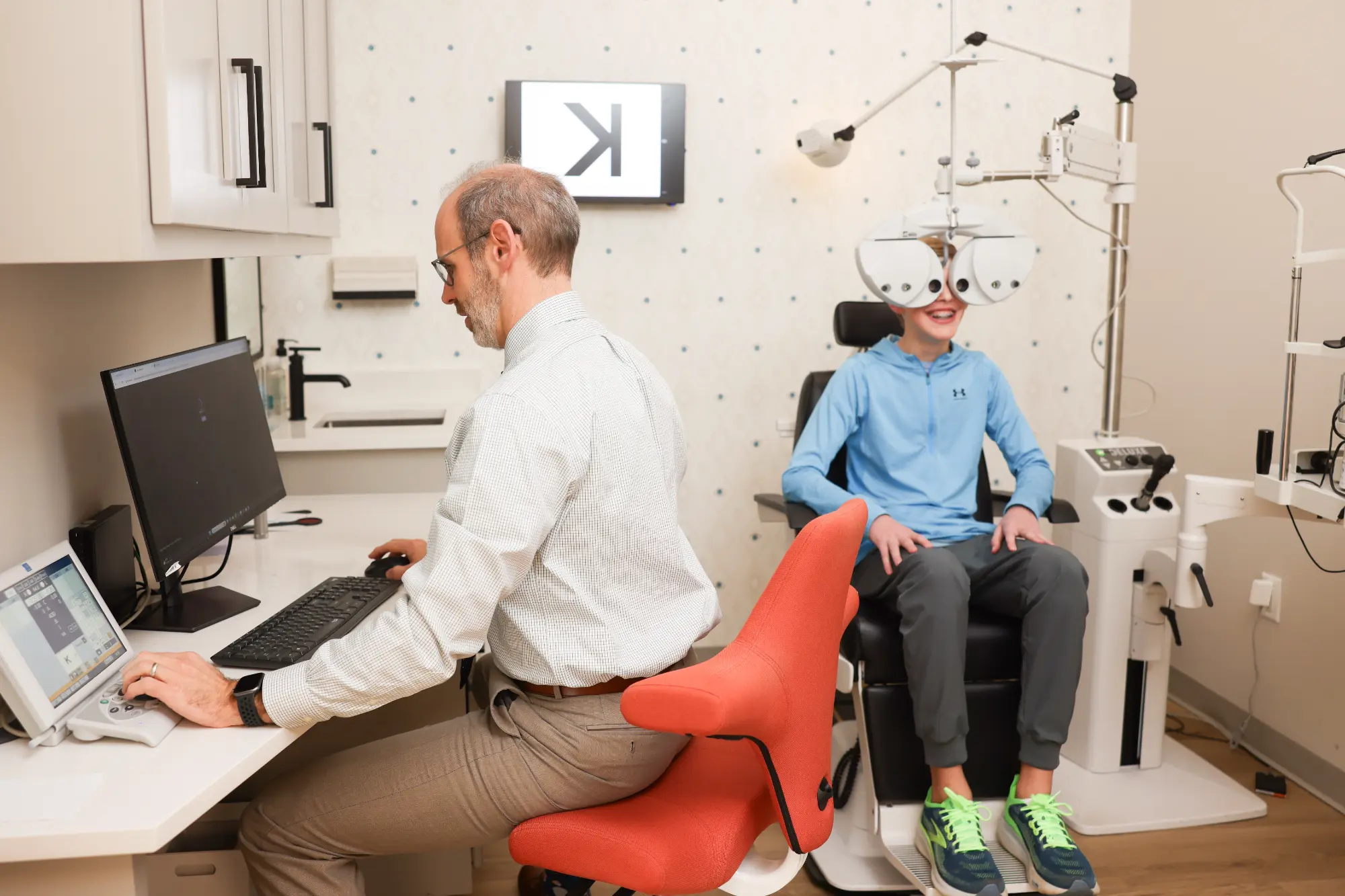 Optometrist Jon Lutz works at his desk while a smiling boy receives an eye exam using a phoropter in the office.