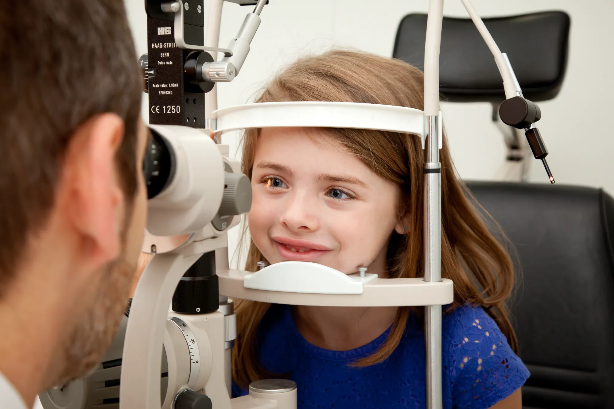An optometrist examines a young girl's eye using specialized equipment. The girl is calm and slightly smiling during the procedure.