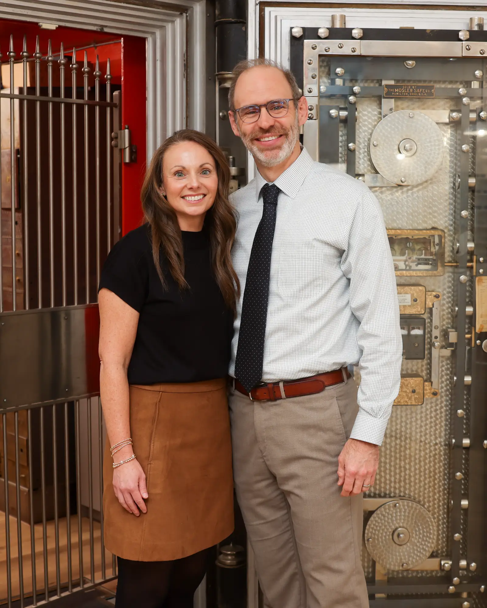 Owners Jon and Janelle Lutz stand smiling warmly in their office against the backdrop of a large, antique bank vault door.