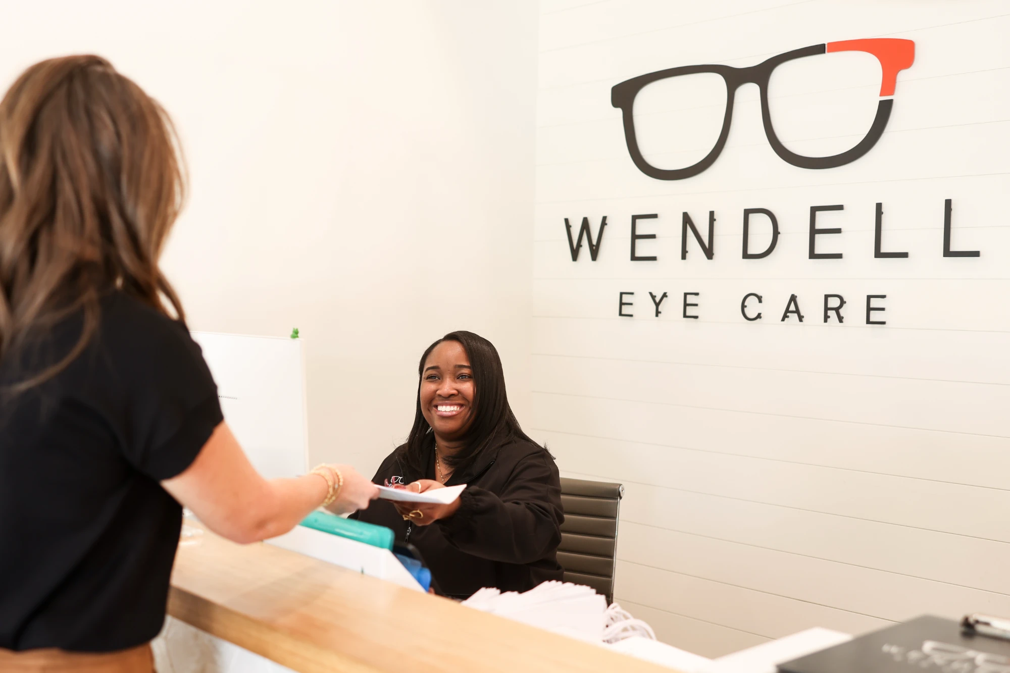 Patient Care Coordinator Myah Johnson smiles brightly while handing a patient paperwork across the reception desk, with the Wendell Eye Care sign visible behind her.
