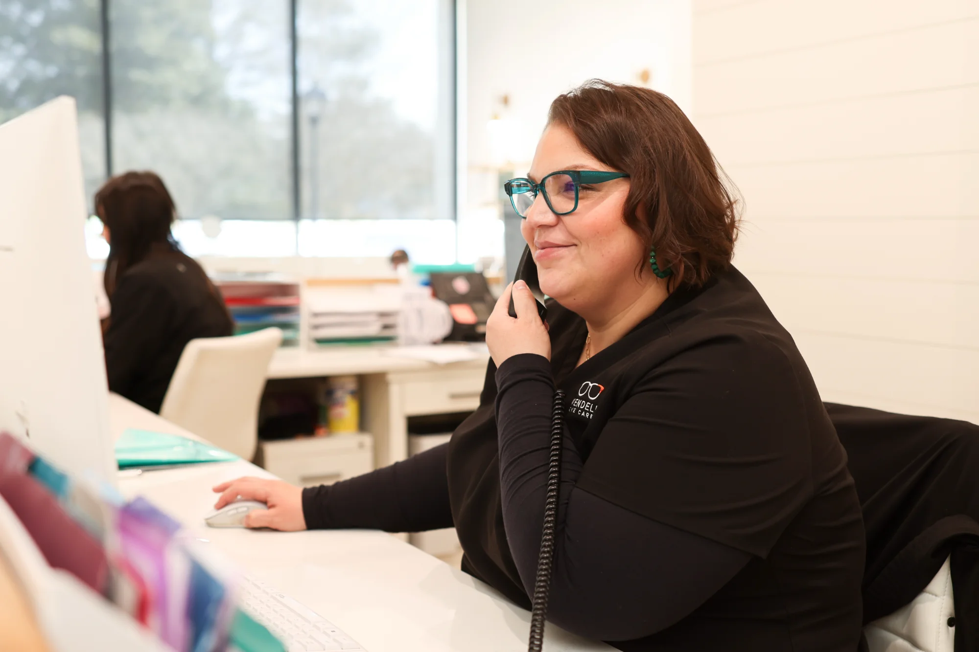 Patient Care Coordinator Sara Marquez, wearing teal glasses, smiles while speaking on the phone and using the computer mouse at the front desk of the office.