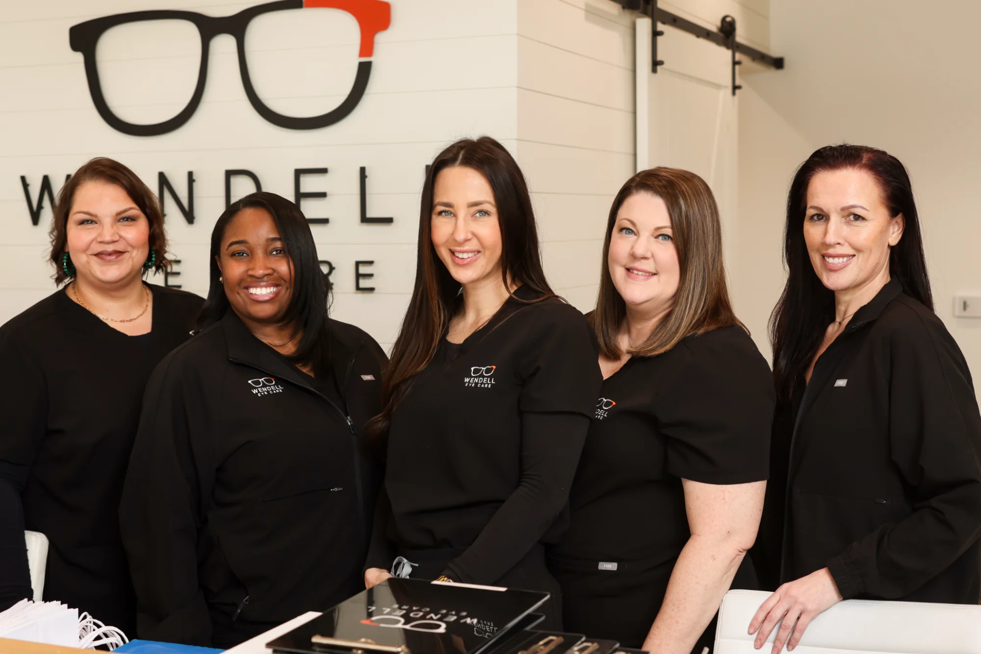 Five Patient Care Coordinators in black scrubs stand smiling confidently at the front desk of Wendell Eye Care, with the large business logo behind them.