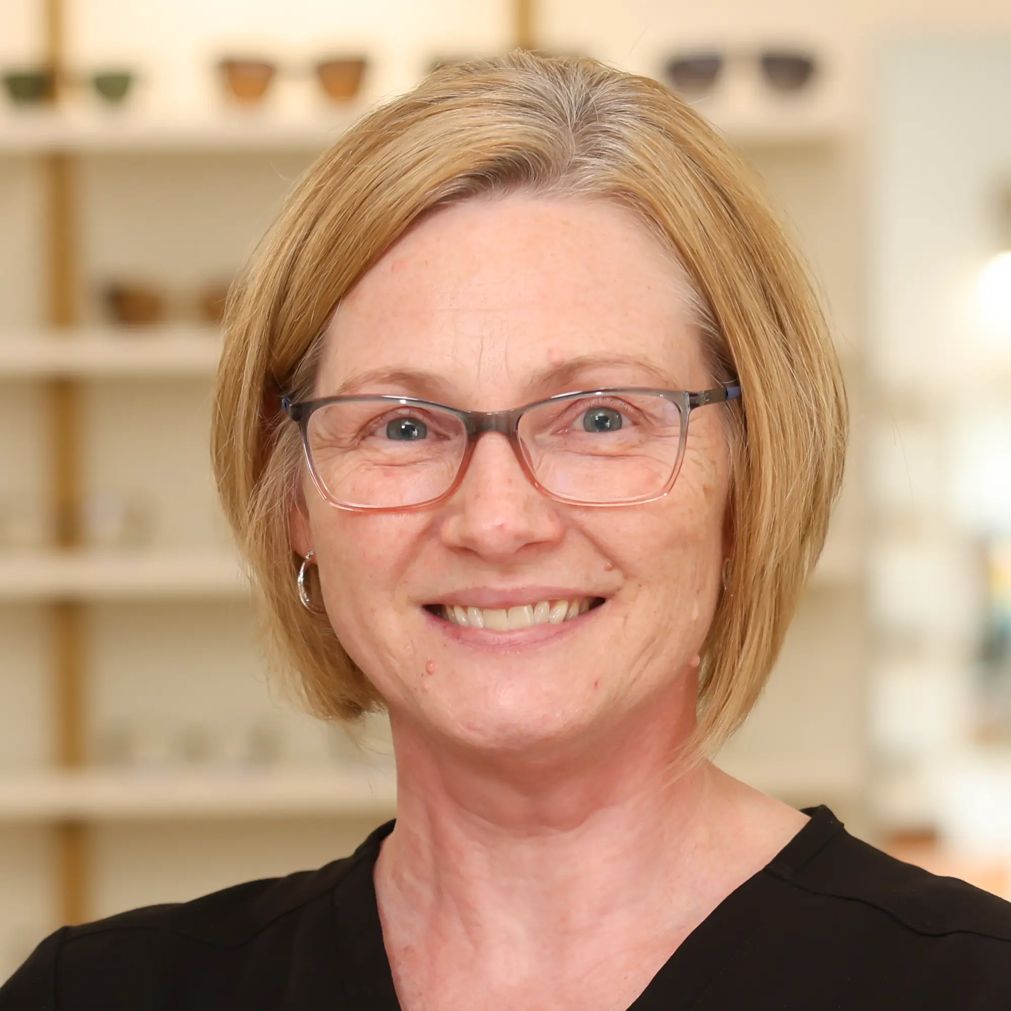 Optometric Technician Crystal Clark, with blonde hair and glasses, smiles brightly in front of a blurred eyewear display.