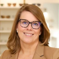 Dr. Holly Bennett, an optometrist, smiles while wearing a tan blazer and large tortoise-shell glasses in her brightly lit office.