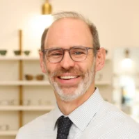 A professional portrait of Dr. Jon Lutz, an optometrist, smiling widely. He wears a checkered shirt, dark tie, and glasses in front of display shelving.