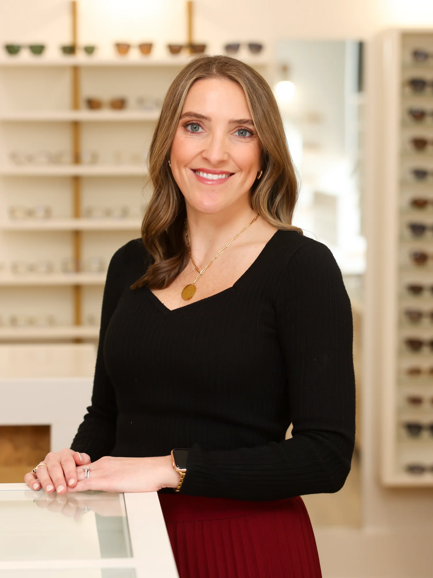 Doctor Rachel Vick, an optometrist, smiles brightly, standing behind a white counter in a well-lit optical office with shelves of glasses displayed.