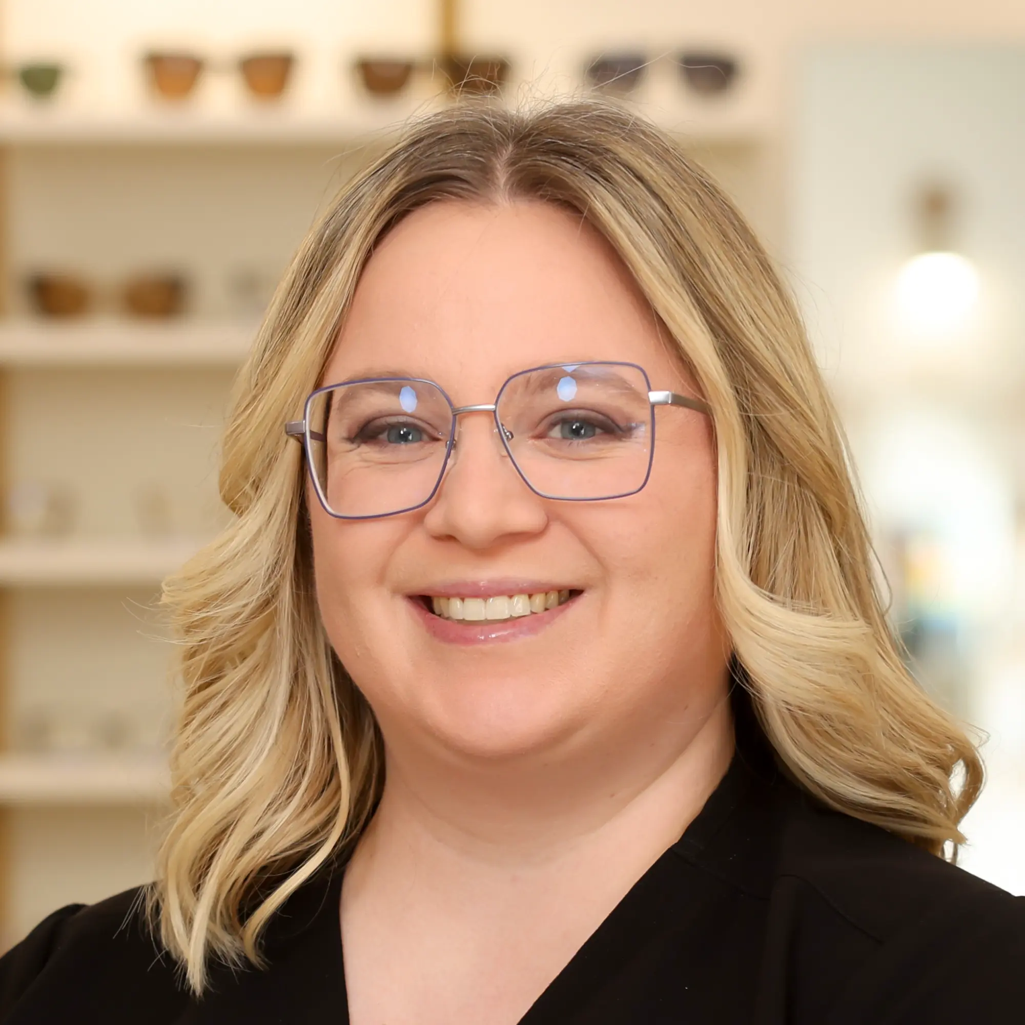 A smiling Emily Kaczynski, Optometric Technician, with blonde hair and square glasses, stands in the store showroom.