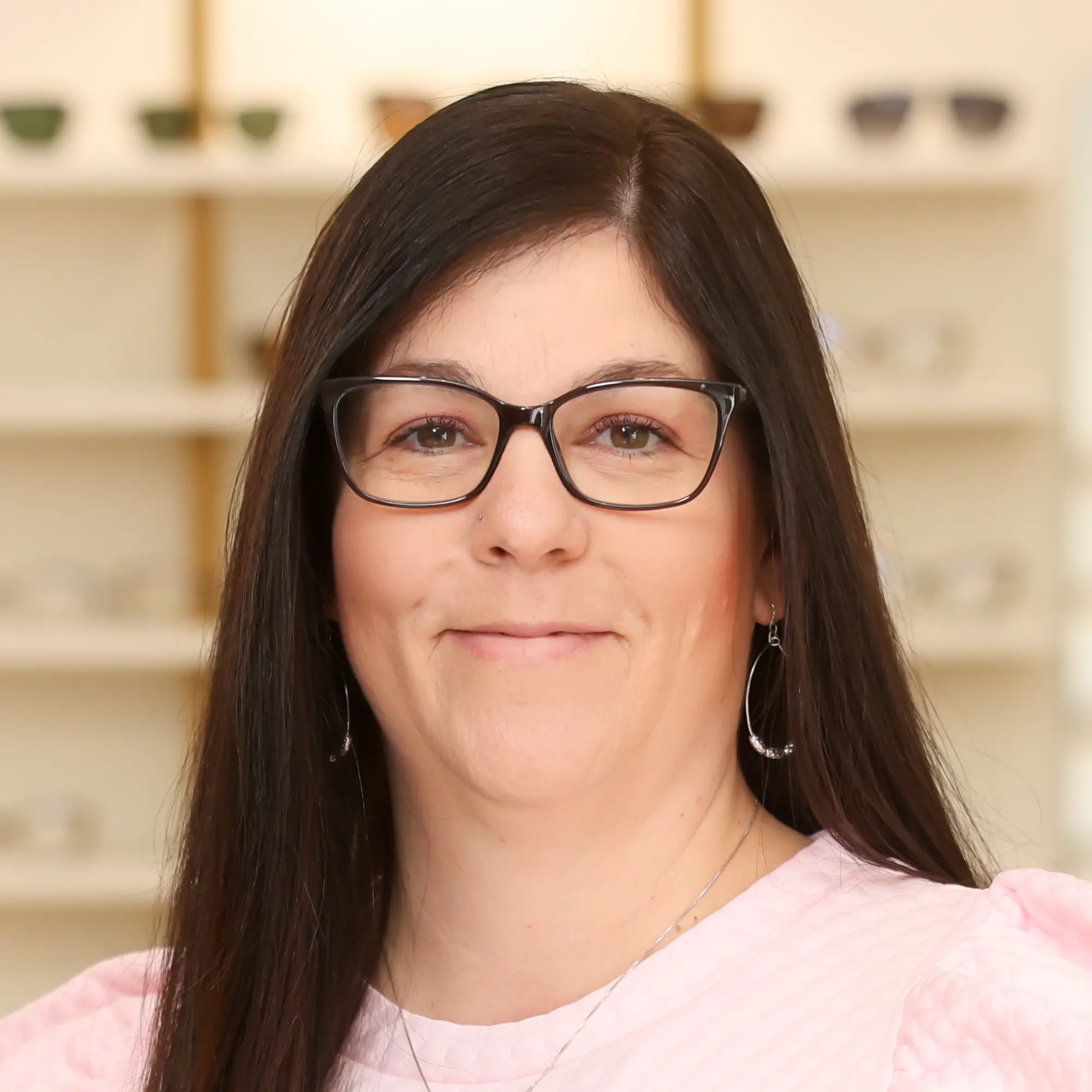 Holly Soles, Optometric Technician, wearing black glasses and a pink top, smiles against a backdrop of optical display shelves.