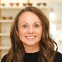 Janelle Lutz, the Office Manager, smiles warmly. She has long brown hair and green eyes, standing in the bright optometrist office near display frames.
