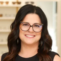 Kelly Metz, an optical apprentice, smiles brightly. She is wearing large tortoise-shell glasses, gold hoop earrings, and has long dark hair.