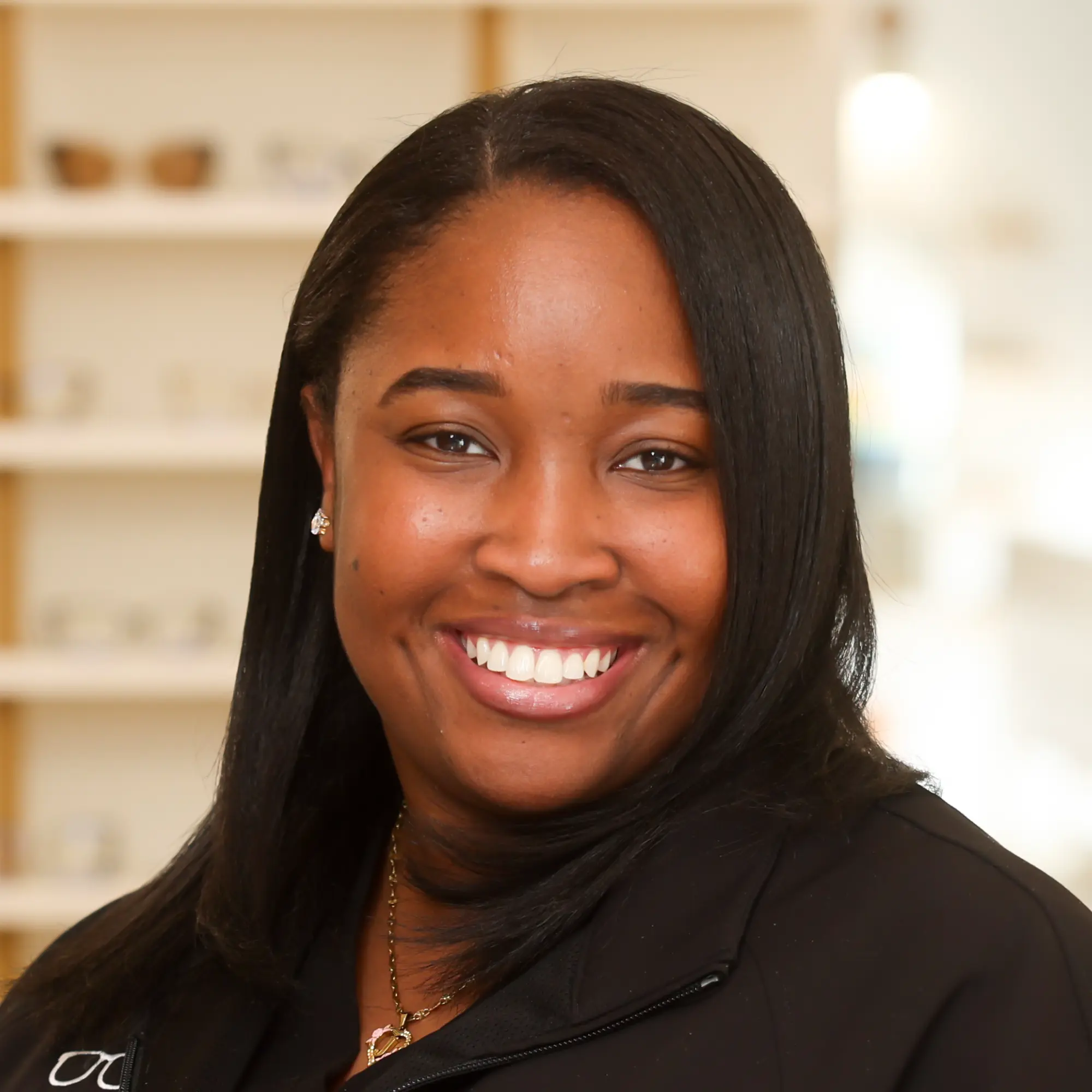 A friendly portrait of Myah Johnson, Patient Care Coordinator, smiling broadly, wearing a black jacket and gold necklace in a bright optical setting.