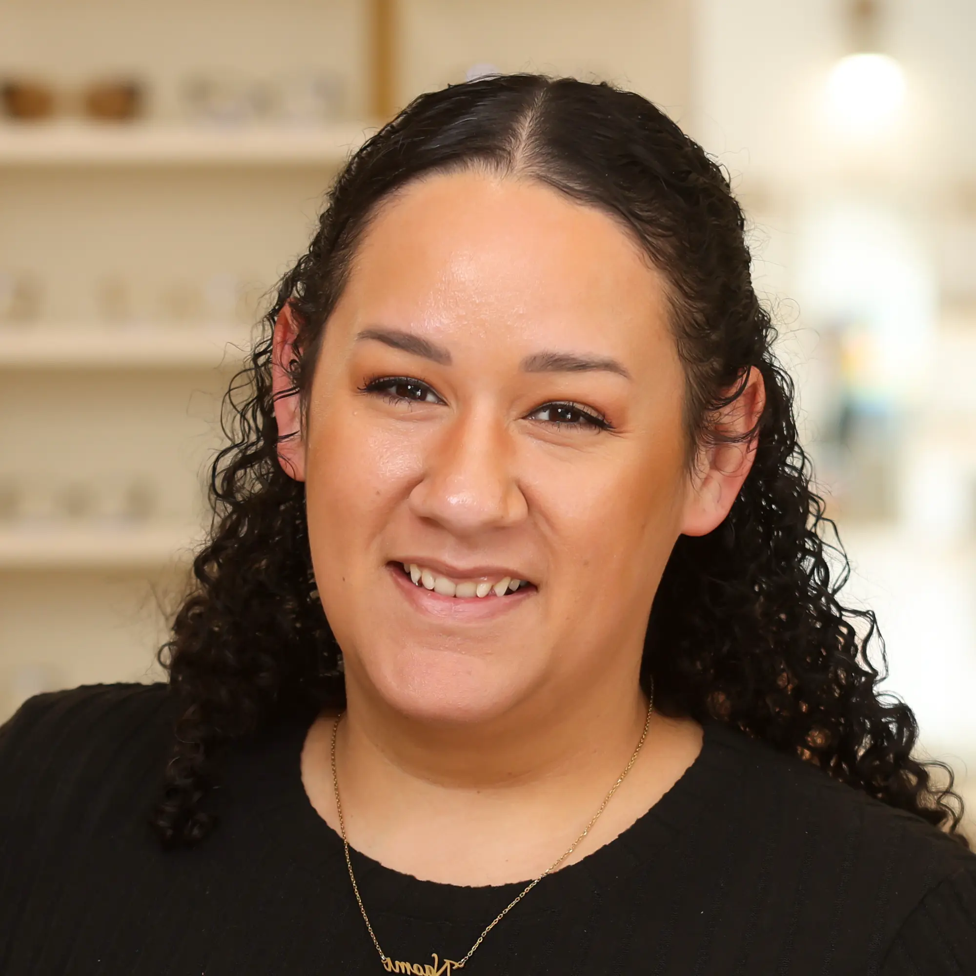 A professional woman with dark curly hair and a gold pendant necklace smiles directly at the camera in a bright, blurred office interior.