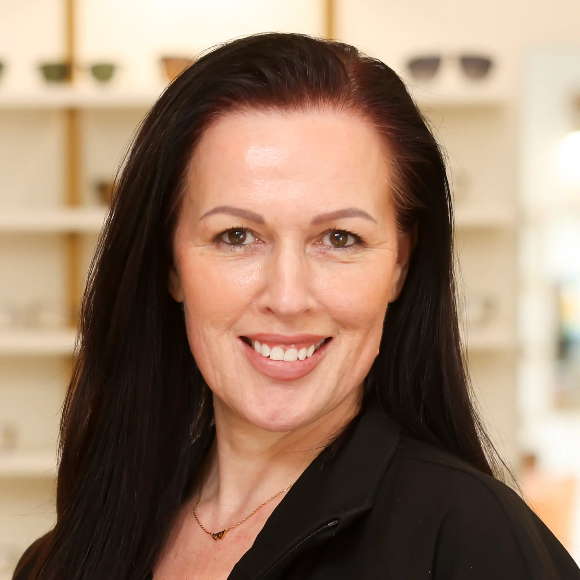 Nikki Winterholler, the Patient Care Coordinator, stands for a professional portrait. She has long dark hair, wears a gold necklace, and smiles warmly at the viewer.