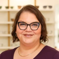 Portrait of Sara Marquez, a Patient Care Coordinator, smiling warmly. She wears stylish black cat-eye glasses, a dark purple top, and gold jewelry in an optical setting.
