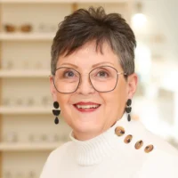 Susan Murphy, a licensed optician, stands in her office, smiling brightly. She wears black geometric glasses and a white sweater with decorative buttons.