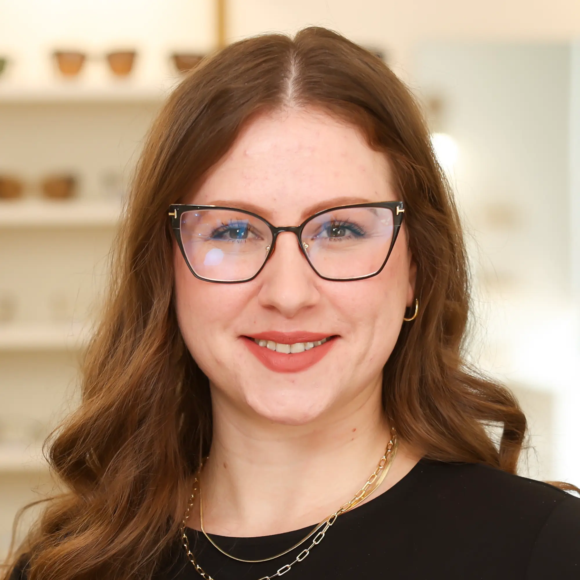 Optical Technician Tiffany Reed smiles for a portrait. She has long brown hair and wears black cat-eye glasses and layered gold necklaces.