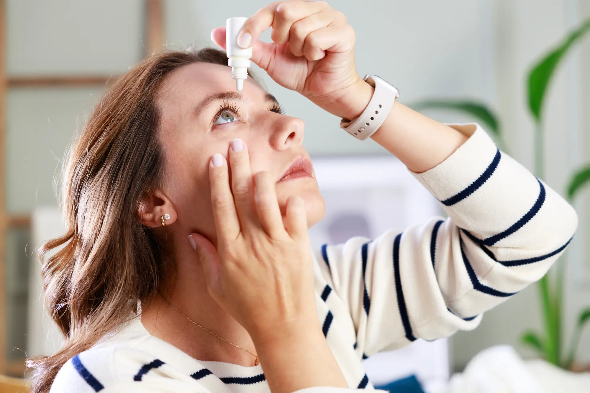 A woman in a striped sweater looks up, concentrating as she administers eye drops into her eye with a steady hand.