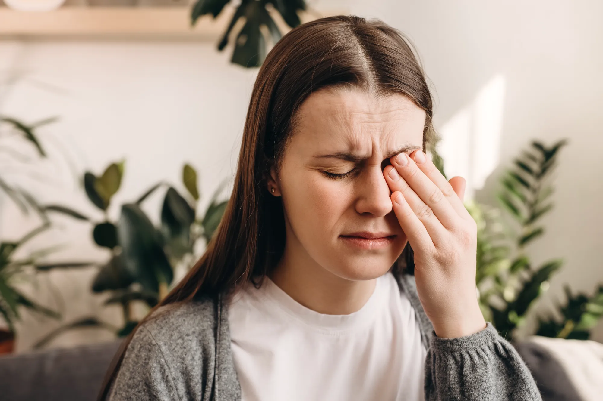 A woman indoors rubs her closed right eye with her hand, wearing a grey sweater and showing a strained expression, suggesting irritation or pain.