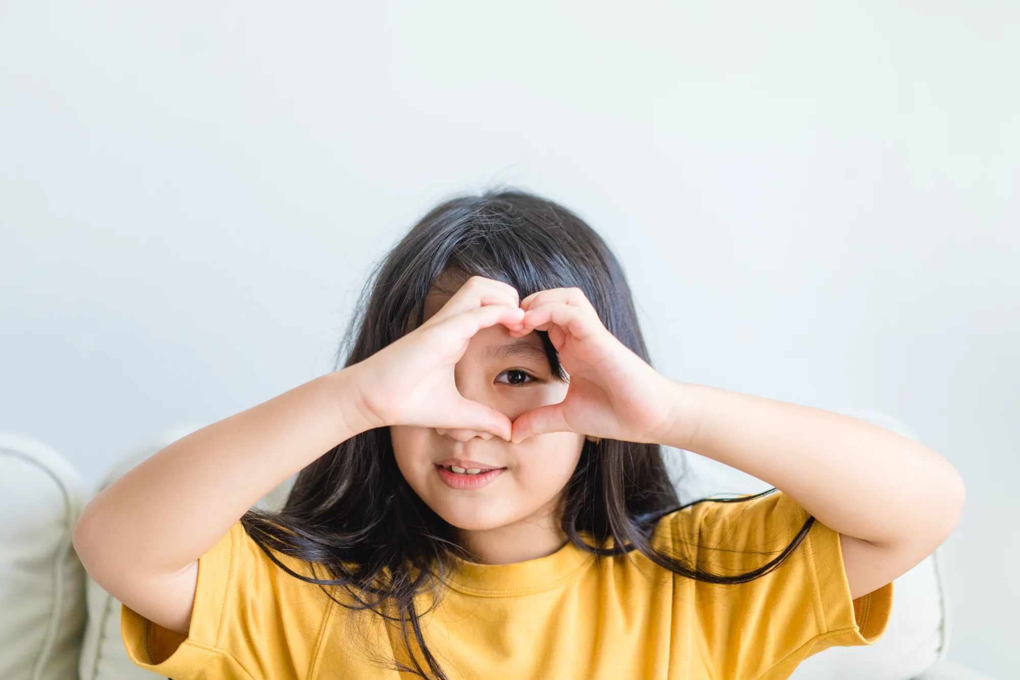 A smiling girl in a yellow shirt holds her hands around her eyes to form a heart shape against a light background.