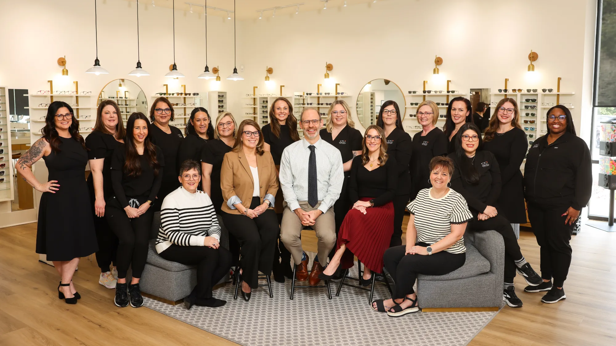 A large team of staff members, all smiling, pose in an optical shop, standing and seated among shelves displaying various eyeglasses and sunglasses.