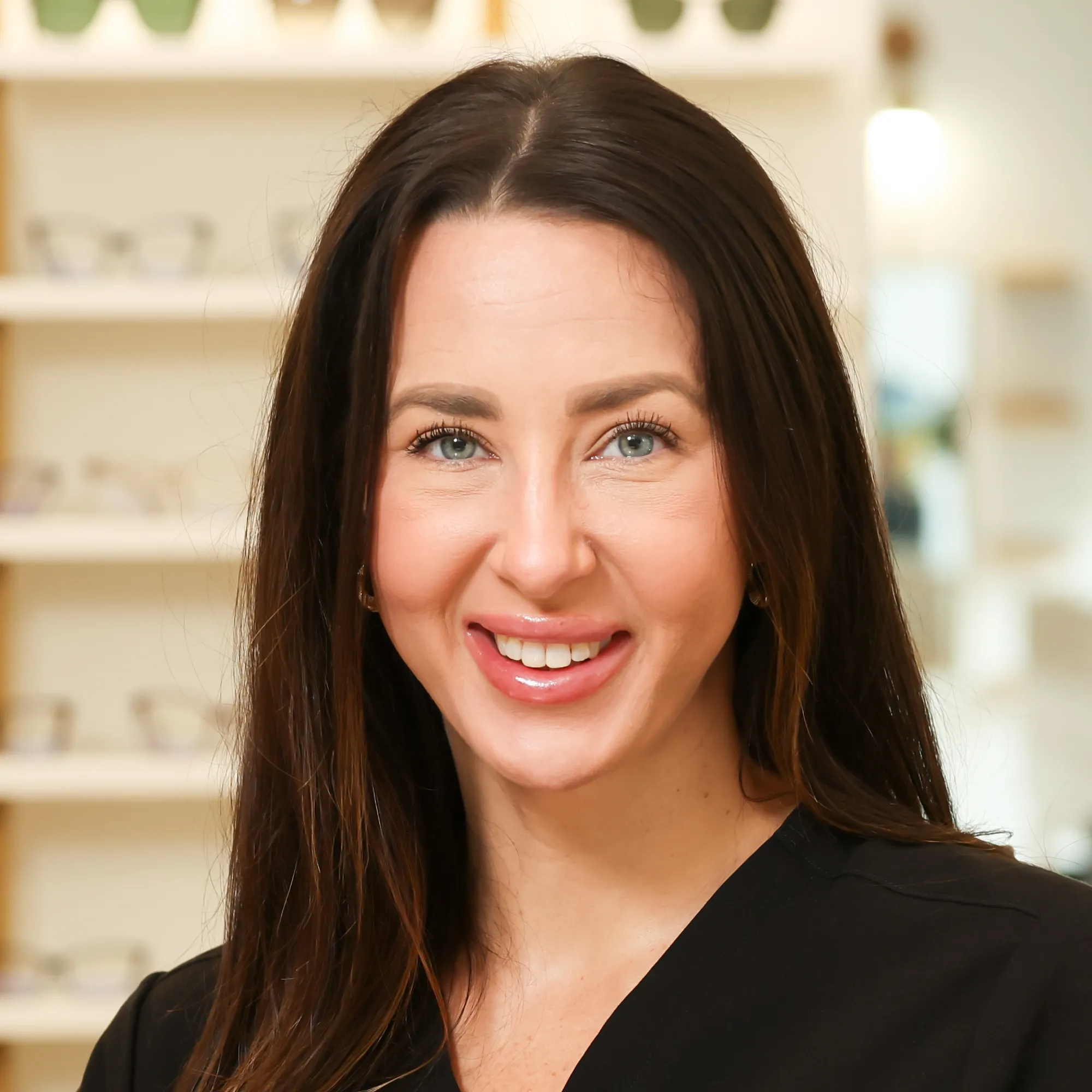 Patient Care Coordinator Chelsea McGuinness, with long brown hair, smiles brightly while standing in front of a display of eyeglasses.