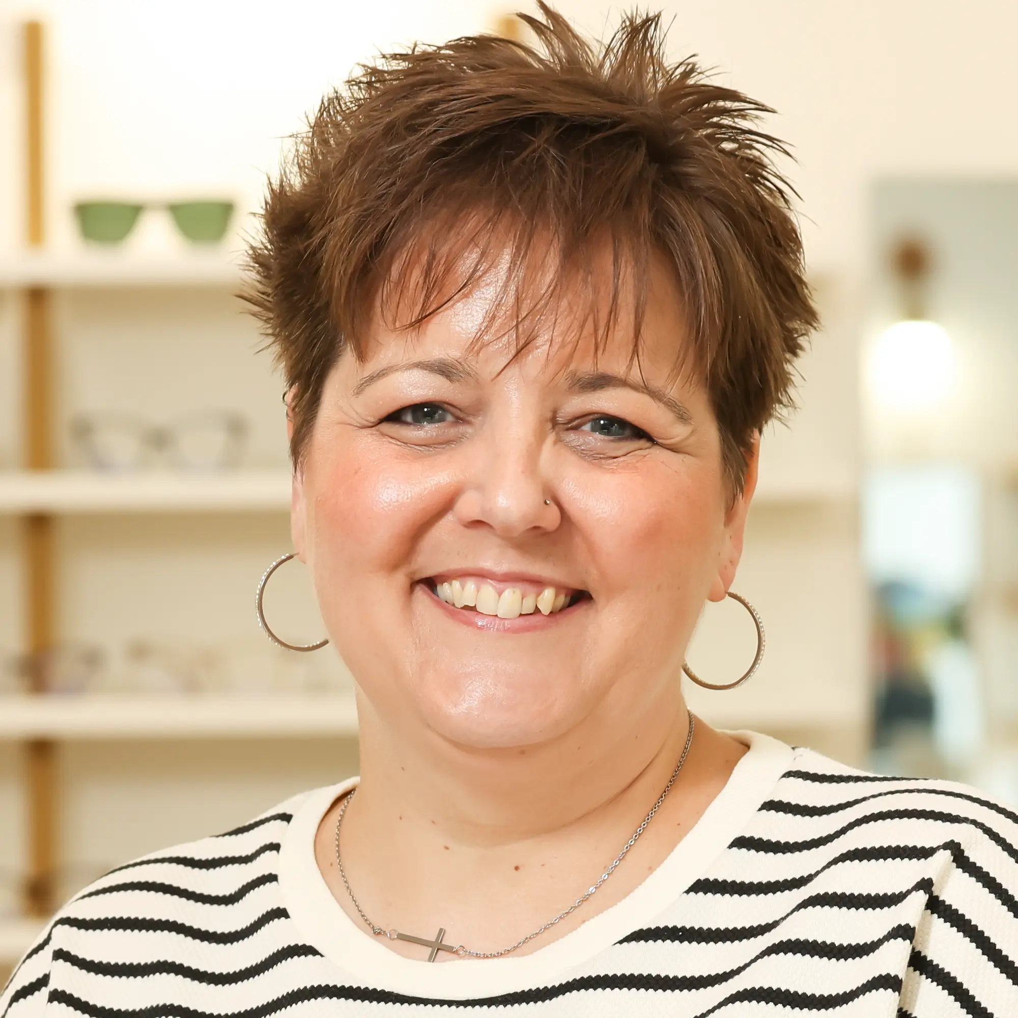 Jodie Priemer, an Optician, with short brown hair and hoop earrings, smiles warmly in the optical clinic showroom.