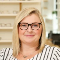 Nichole Honeycutt, an Optometric Technician, smiles in an optical office setting. She has blonde hair, wears tortoise-shell glasses, a striped shirt, and a nose ring.