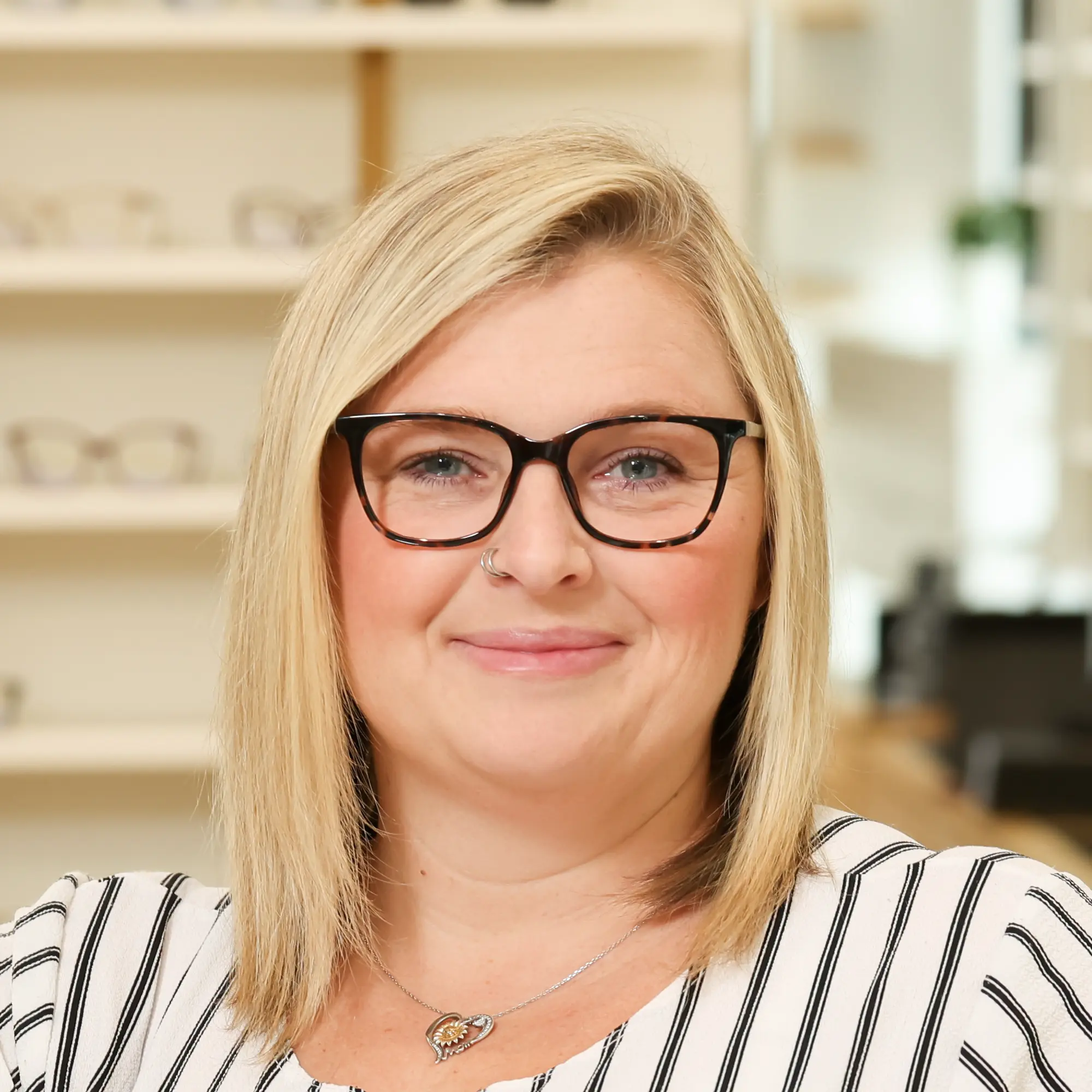 Nichole Honeycutt, an Optometric Technician, smiles in an optical office setting. She has blonde hair, wears tortoise-shell glasses, a striped shirt, and a nose ring.